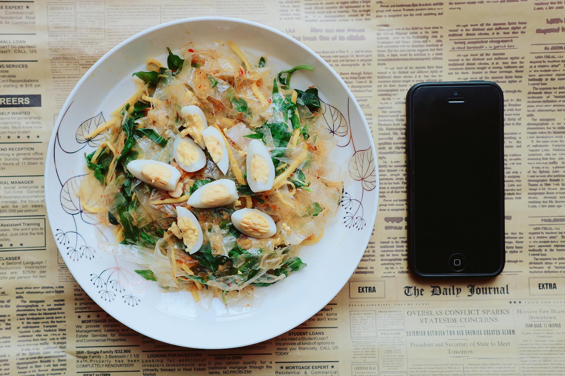 Top view of an Asian dish with boiled eggs and greens beside a smartphone on a newspaper background.